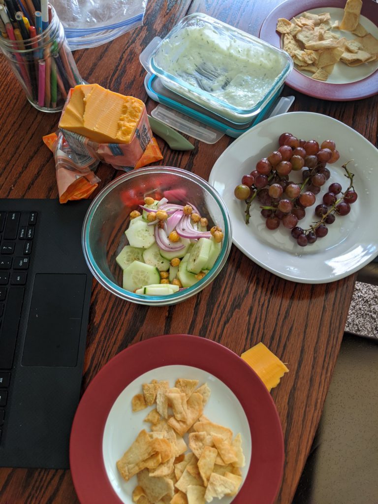 2 plates with broken pita chips and a block of cheese in wrapper, half eaten container of salad, sauce, and some grapes on a workstation